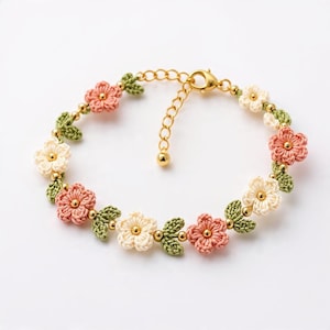 May include: A delicate crochet bracelet featuring a floral design. The bracelet showcases pink, cream, and green crocheted flowers and leaves, accented with gold-colored beads and a clasp. The bracelet is approximately 7 inches long.