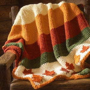 May include: A handmade crochet blanket draped over a wooden chair. The blanket features horizontal stripes of cream, orange, rust, green, and white, with appliqued orange leaf details. The blanket is a cozy, autumn-themed home decor item.