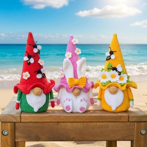 Pode incluir: Três gnomos de crochê feitos à mão. Um tem um chapéu vermelho com abelhas e flores brancas, outro um chapéu rosa com orelhas de coelho e o terceiro um chapéu amarelo com abelhas e margaridas. Estão numa mesa de madeira numa praia.