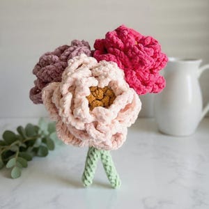 May include: A bouquet of handmade crochet flowers in shades of pink, mauve, and yellow. The flowers have a textured, plush appearance and are arranged with green stems. A white pitcher is in the background.