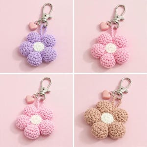May include: Four crochet flower keychains in various colors: lavender, pink, light pink, and tan. Each flower has a white center and a small heart charm. The keychains have silver-colored clasps and are displayed on a pink background.