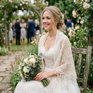 Peut inclure: Une mariée en robe de mariée en dentelle blanche et châle est assise sur un banc en bois, tenant un bouquet de fleurs blanches et de verdure. L'arrière-plan présente un chemin pavé, des rosiers et des invités.