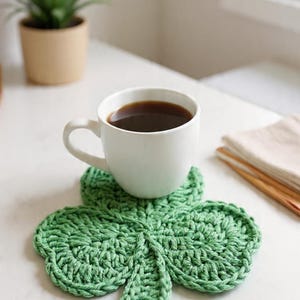 May include: A white ceramic mug filled with dark coffee sits on a green, crocheted shamrock-shaped coaster. The coaster is on a white surface, with a small potted plant and a window in the background. A stack of folded napkins and wooden sticks are also visible.