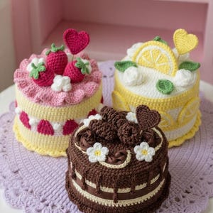 Può includere: Tre torte all'uncinetto in rosa, giallo e marrone, ognuna con decorazioni uniche. La torta rosa ha fragole e un cuore, la torta gialla presenta fette di limone e la torta marrone è ricoperta di riccioli di cioccolato e fiori. Sono su una tovaglietta all'uncinetto lavanda.