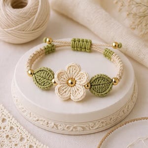 Puede incluir: Una pulsera de macramé hecha a mano con una flor color crema y hojas verdes. La pulsera tiene cuentas y detalles dorados. El cierre ajustable es de macramé verde. La pulsera se exhibe sobre una superficie blanca decorativa.