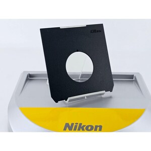 May include: A black Nikon camera filter with a circular opening is displayed on a silver and yellow stand. The Nikon logo is visible on the filter and the stand. The filter is rectangular with a textured surface.
