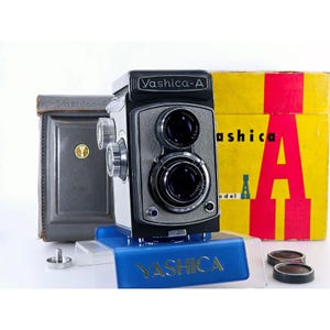 May include: A black and gray Yashica-A twin-lens reflex camera with two circular lenses, silver adjustment knobs, and a Copal shutter. It is displayed with a gray leather case, a yellow box with red text, and two small circular lens caps.