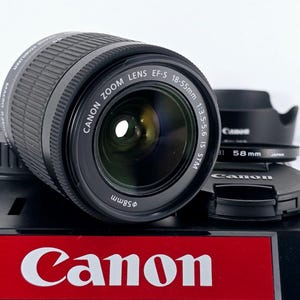 Può includere: Un obiettivo Canon Zoom EF-S 18-55mm 1:3.5-5.6 IS STM, con un diametro filtro di 58mm, è in mostra. È accompagnato da un tappo e un paraluce. Il logo Canon è visibile in rosso e bianco.