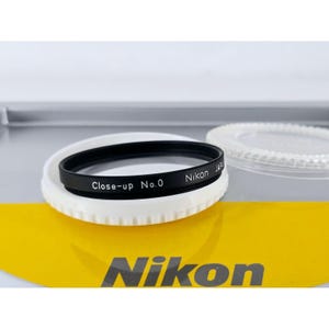 Könnte beinhalten: Ein schwarzer, kreisförmiger Nikon Close-up No.0 Objektivfilter mit weißem Text auf der Seite. Der Filter liegt auf einem weißen Kunststoffring, und ein separater, durchsichtiger Kunststoff-Objektivdeckel ist im Hintergrund zu sehen.