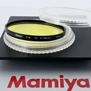 May include: A circular yellow Mamiya camera lens filter with a black metal frame. The rim is printed with the text Mamiya 77ø Y2 S Y48.2 C JAPAN. The filter rests on a clear plastic circular case lid, which is placed on a black square base.