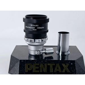 May include: A black and silver Asahi Microscope Adapter with Japan printed on the side. The adapter features a cylindrical metal body with a knurled adjustment knob and a separate silver metal tube component, all displayed on a black base.