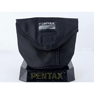 May include: A black, textured fabric camera pouch with a fold-over flap and a rectangular patch featuring the white text PENTAX. The pouch is positioned on a black, speckled, angular base that also displays the word PENTAX in gold-colored lettering.