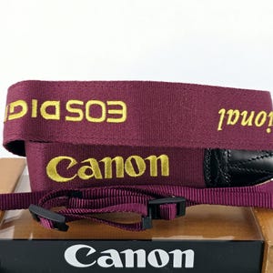 May include: A burgundy camera strap featuring the yellow embroidered text Canon and EOS DIGITAL. The strap includes black leather-like end attachments and adjustable black plastic buckles, designed for securing a camera during use.