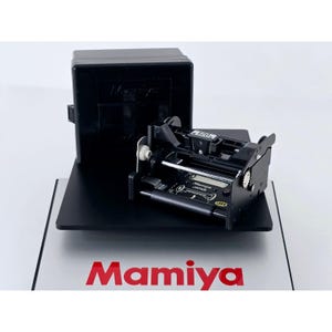 May include: A black Mamiya 120 film insert for a medium format camera, featuring a metal and plastic frame with internal rollers and gears. The insert is positioned in front of its black storage case, with the Mamiya logo printed on the base.