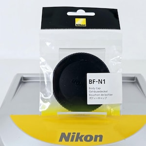 May include: A black Nikon BF-N1 body cap for a camera, enclosed in a clear plastic retail package with a yellow and black header. The label on the front of the packaging displays the text BF-N1, Body Cap, Gehäusedeckel, Bouchon de boîtier.