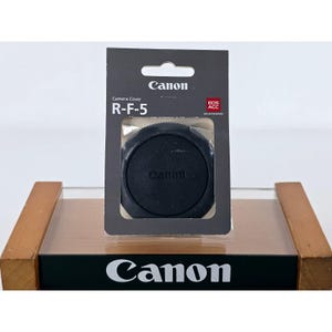 Op de afbeelding: Een zwarte Canon R-F-5 camerabody-dop in de originele donkergrijze verpakking. De verpakking bevat het Canon-logo, de tekst Camera Cover R-F-5 en een rood EOS ACC-vierkant. De ronde zwarte dop is zichtbaar door een uitgesneden venster.