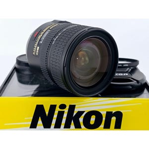 May include: A black Nikon AF-S Nikkor 24-120mm 1:3.5-5.6 G ED VR camera lens with a textured zoom ring and focus ring. The lens is positioned on a yellow box with the black Nikon logo printed on the front, accompanied by a black lens cap.