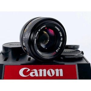 May include: A black Canon FD 55mm 1:1.2 S.S.C. camera lens with a textured focus ring and orange distance markings. The lens features the text 'CANON LENS FD 55mm 1:1.2 S.S.C.' and 'LENS MADE IN JAPAN' printed on the front element housing.