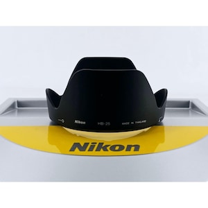 May include: A black Nikon HB-25 camera lens hood with a petal-shaped design. The exterior features white printed text that reads Nikon, HB-25, and MADE IN THAILAND, along with alignment indicators. The lens hood is positioned on a yellow and gray surface.
