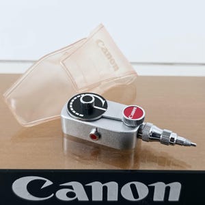 May include: A silver Canon camera accessory featuring a black circular dial with white markings, a red button with the Canon logo, and a small red side button. It is accompanied by a translucent plastic case with the Canon logo embossed on it.
