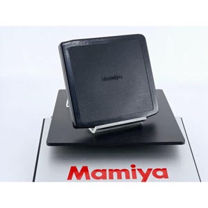 May include: A black square Mamiya camera film back cover with rounded corners, positioned on a clear acrylic stand. Below it, a silver rectangular plate features the red Mamiya logo printed in a bold, sans-serif font on its surface.