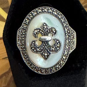 May include: An oval ring featuring a central white mother-of-pearl inlay decorated with a silver fleur-de-lis emblem. The border of the ring is encrusted with small, dark, faceted marcasite stones, creating a textured metallic frame around the center.