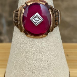 Könnte beinhalten: Ein Goldring mit einem großen, ovalen, dunkelroten Cabochon-Stein in der Mitte. Eine kleine, quadratische, silberfarbene Fassung mit einem einzelnen klaren Stein befindet sich direkt in der Mitte des roten Cabochons.