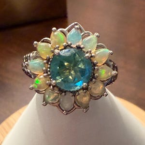 May include: A silver ring featuring a large, round, faceted blue center stone surrounded by a halo of twelve pear-shaped opals. The metal band has a textured, intricate pattern along the sides, and the setting creates a floral-like arrangement.