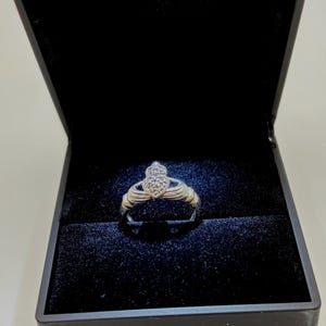 May include: A silver Claddagh ring featuring a heart-shaped center set with small clear stones, held by two hands, resting inside a black velvet-lined jewelry box with a hinged lid that is open to display the ring clearly against the dark fabric.