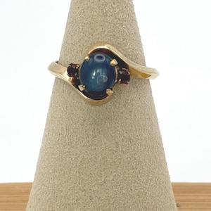 Puede incluir: Un anillo de oro con una piedra central ovalada de color azul oscuro tallada en cabujón. El aro presenta un diseño cruzado con una pequeña piedra preciosa facetada de color rojo oscuro a cada lado, sujeta por garras de oro en el aro.