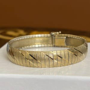 May include: A gold-tone metal bracelet featuring a wide, flat, flexible band composed of interlocking rectangular links. The surface of the links displays a textured, diagonal striped pattern, and the bracelet includes a secure box clasp closure.