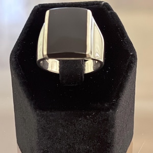 May include: A silver-toned ring featuring a smooth, rectangular black stone inlay set into the center of the band. The metal band has a polished, reflective finish and a wide, solid construction that tapers slightly toward the bottom of the ring.
