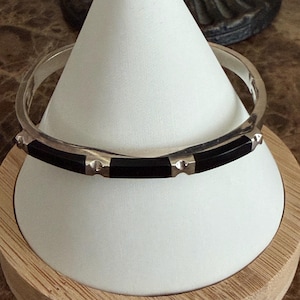 Peut inclure: Un bracelet jonc en argent composé de segments alternés en argent poli et en incrustation noire lisse. Le design comprend de petits accents argentés arrondis placés entre chaque section noire, formant un anneau circulaire continu.