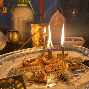 May include: A spiritual altar with burning candles, incense, and a small paper with text. The altar includes a silver tray with salt, herbs, and lemon slices. A white vase with green plants is in the background.
