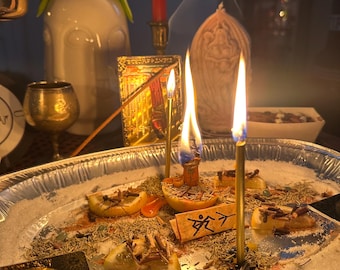 Pode incluir: Um altar espiritual com velas acesas, incenso e um pequeno papel com texto. O altar inclui uma bandeja de prata com sal, ervas e rodelas de limão. Um vaso branco com plantas verdes está ao fundo.