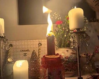Pode incluir: Uma vela vermelha acesa num frasco de vidro com uma carta de tarot que diz "THE LOVERS". Uma vela branca queima num suporte decorativo. Um vaso de flores e uma pequena árvore de Natal também são visíveis.