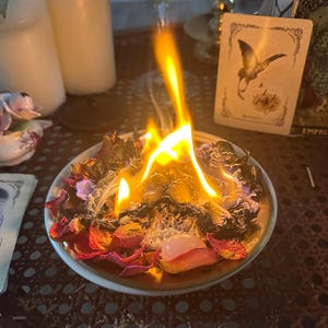 May include: A burning arrangement of dried rose petals and herbs in a small, gold-rimmed dish. Flames rise from the center, with a rose quartz crystal visible. Tarot cards and candles are in the background, suggesting a spiritual or ritualistic theme.