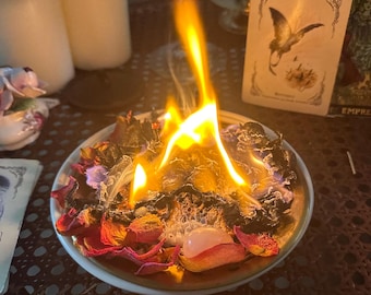 May include: A burning arrangement of dried rose petals and herbs in a small, gold-rimmed dish. Flames rise from the center, with a rose quartz crystal visible. Tarot cards and candles are in the background, suggesting a spiritual or ritualistic theme.