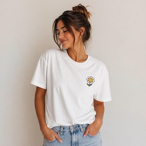 May include: White t-shirt with a small embroidered yellow daisy with a smiling face. The daisy has a black outline and green stem. The shirt is paired with blue jeans.