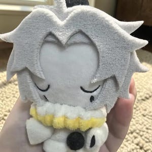 Puede incluir: Una mu&ntilde;eca de peluche con pelo gris y una cara blanca. La mu&ntilde;eca tiene un cuello blanco y amarillo y un bot&oacute;n negro en la parte delantera. La mu&ntilde;eca est&aacute; dise&ntilde;ada para parecerse a un personaje de videojuego o anime.