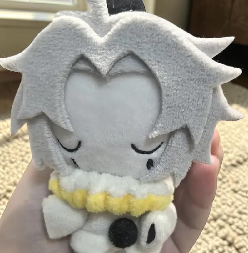 Puede incluir: Una mu&ntilde;eca de peluche con pelo gris y una cara blanca. La mu&ntilde;eca tiene un cuello blanco y amarillo y un bot&oacute;n negro en la parte delantera. La mu&ntilde;eca est&aacute; dise&ntilde;ada para parecerse a un personaje de videojuego o anime.