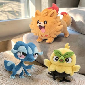 May include: Three plush toys of cartoon creatures. The top toy is a light orange dog-like creature with a red flame-like design. Below is a blue lizard-like creature. The third toy is a yellow owl-like creature with green accents.
