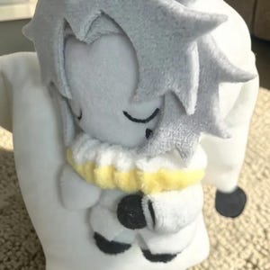 Puede incluir: Una mu&ntilde;eca de peluche en posici&oacute;n de dormir, con cuerpo blanco y cabeza gris claro con pelo puntiagudo. La mu&ntilde;eca tiene los ojos cerrados y un cuello amarillo y blanco. La mu&ntilde;eca sostiene una almohada blanca.