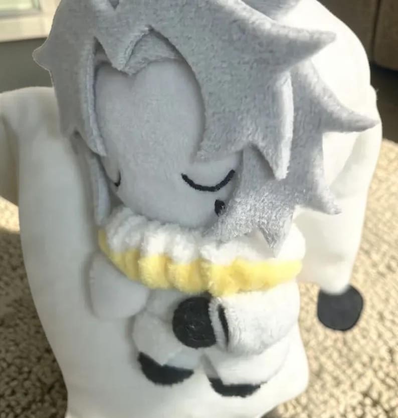 Puede incluir: Una mu&ntilde;eca de peluche en posici&oacute;n de dormir, con cuerpo blanco y cabeza gris claro con pelo puntiagudo. La mu&ntilde;eca tiene los ojos cerrados y un cuello amarillo y blanco. La mu&ntilde;eca sostiene una almohada blanca.