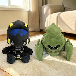 May include: Two plush toys are displayed on a light-coloured rug. One is a black figure with yellow detailing and a blue visor. The other is a green, insect-like creature with large, outstretched arms. Both toys are soft and detailed.