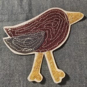 May include: A fabric bird patch featuring a dark maroon body with white stitched contour lines, a grey wing, and a yellow beak and legs. The bird is constructed from layered fabric pieces with visible white thread stitching along the edges.