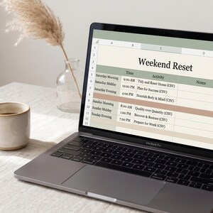 May include: A laptop displaying a "Weekend Reset" schedule, with a coffee mug and decorative plant in the background. The schedule includes time slots and activities for Saturday and Sunday. The laptop is open on a white surface.