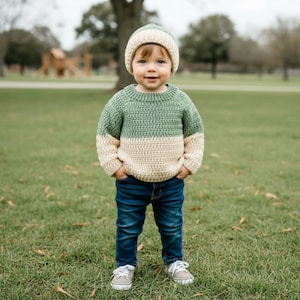 May include: A child wearing a two-toned crochet sweater with a sage green upper section and a cream lower section, paired with a matching cream and sage green crochet beanie, dark blue denim jeans, and light gray canvas sneakers with white laces.