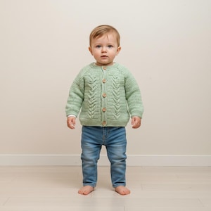 Peut inclure: Un cardigan vert clair pour bébé, tricoté avec un motif en torsade sur le devant. Le cardigan a des boutons en bois et est associé à un jean en denim bleu. Le bébé est pieds nus.