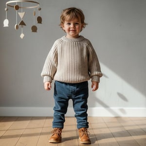 Könnte beinhalten: Ein junges Kind trägt einen beigefarbenen Rippstrickpullover, eine dunkelblaue Hose und braune Lederstiefel. Im Hintergrund hängt ein Holzmobile mit geometrischen Formen. Das Kind steht auf einem Holzboden.